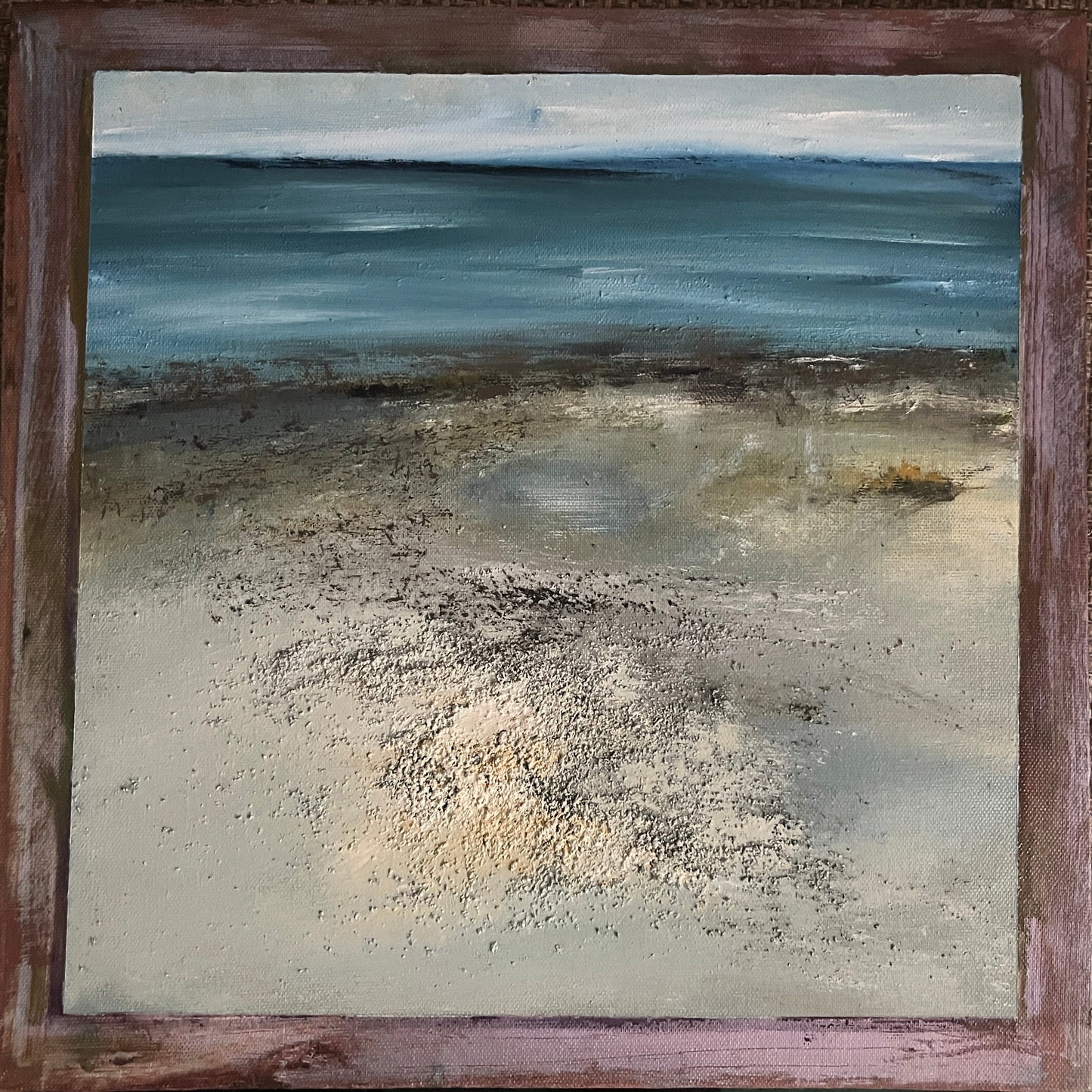 A framed square painting depicting an abstract coastal scene with blue water, a pale sky, and a sandy shore rendered in textured brushstrokes. The artwork is enclosed in a rustic reddish-brown wooden frame.