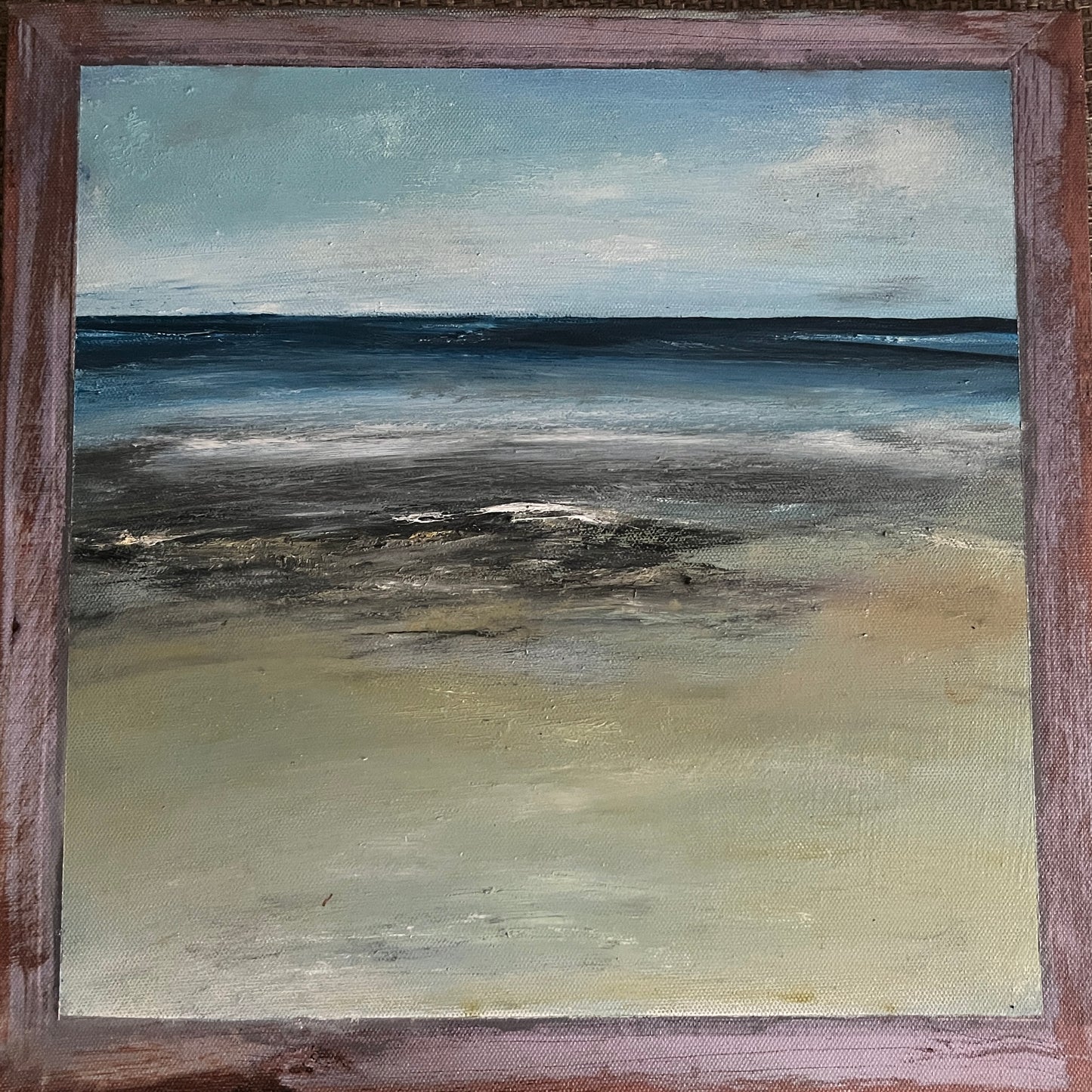 An oil painting of a coastal landscape, depicting a calm sea meeting a sandy shore under a cloudy sky, housed in a rustic reddish-brown wooden frame.